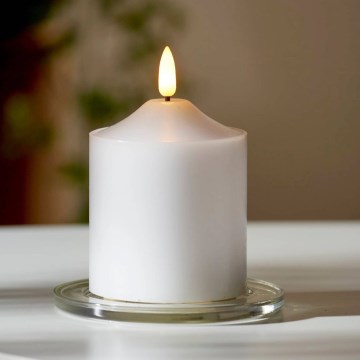Biela elektronická sviečka svieti na bielom pozadí. Sviečka má na vrchu simulovaný plameň.