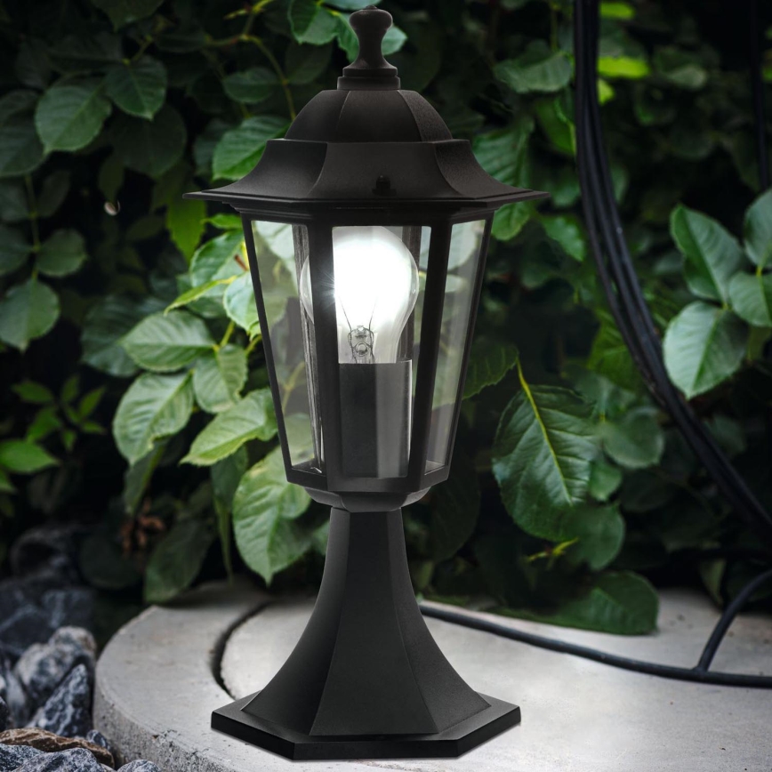 Kovová záhradná lampa svieti, osvetľuje kamenistý okraj a listnatý zelený porast v pozadí.