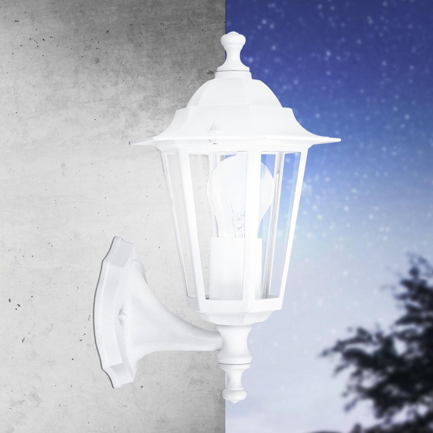 Biela nástenná lampa visí na betónovej stene, osvetľujúca nočné nebo posiate hviezdami. Pozadie obsahuje rozmazaný obrys stromu.