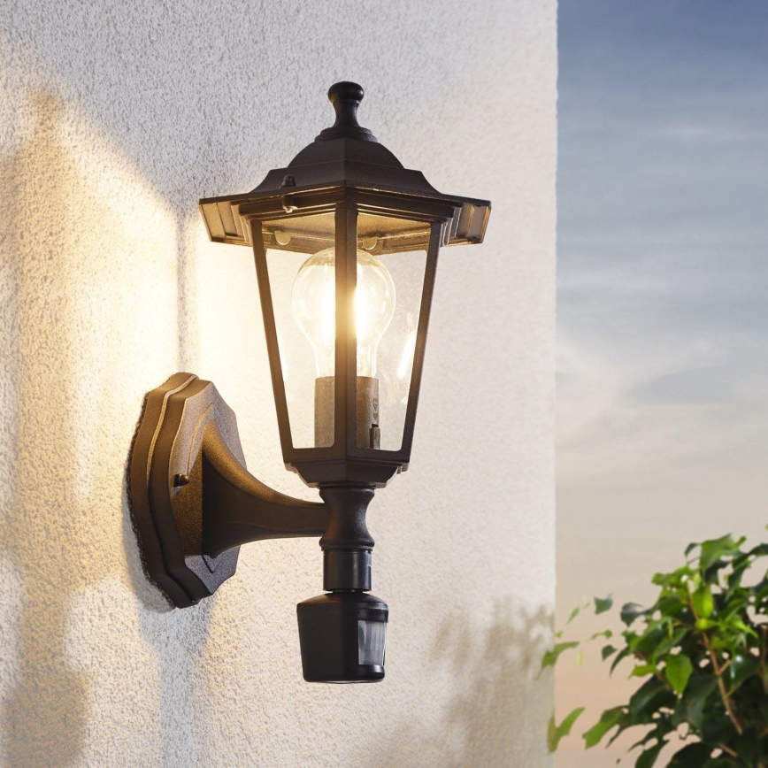 Kovová nástenná lampa svieti na fasáde budovy. Okolo lampy rastliny a otvorené nebo naznačujú vonkajšie prostredie.