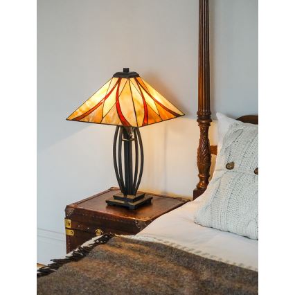 Stolová lampa so štvorcovým tienidlom z farebného skla svieti na nočnom stolíku. Lampa stojí na drevenom nočnom stolíku v spálni pri posteli s čelom.