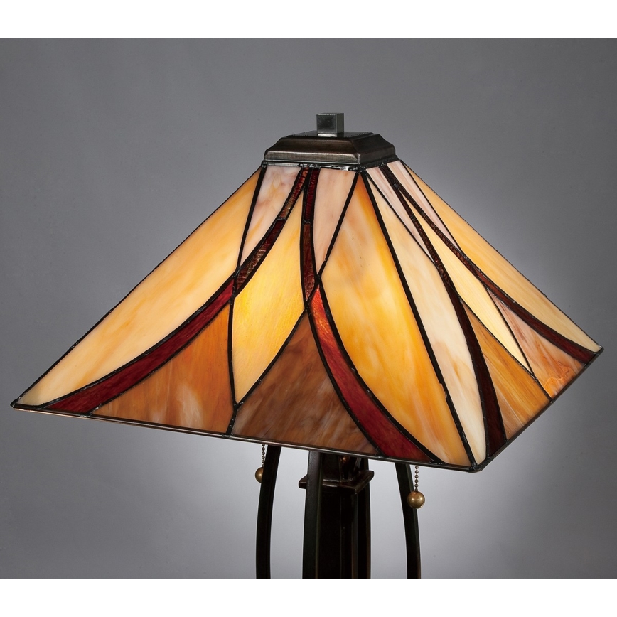 Stolová lampa so štvorcovým tienidlom z farebného skla, v odtieňoch béžovej, hnedej a červenej, stojí na tmavom kovovom podstavci. Tienidlo je zdobené geometrickými vzormi.
