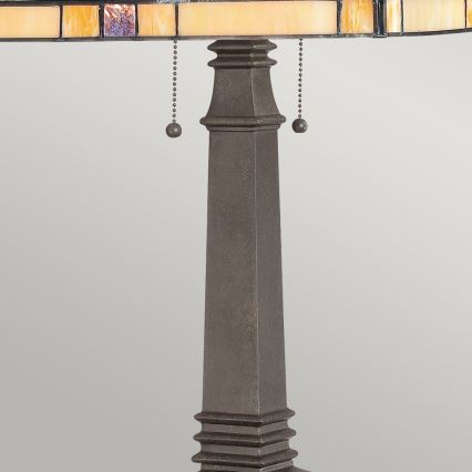 Stolová lampa s tienidlom z farebného skla stojí na tmavosivej kovovej nohe. Tienidlo je zdobené geometrickými vzormi. Noha má zúžený tvar a zakončenie v podobe ozdobných pásov.