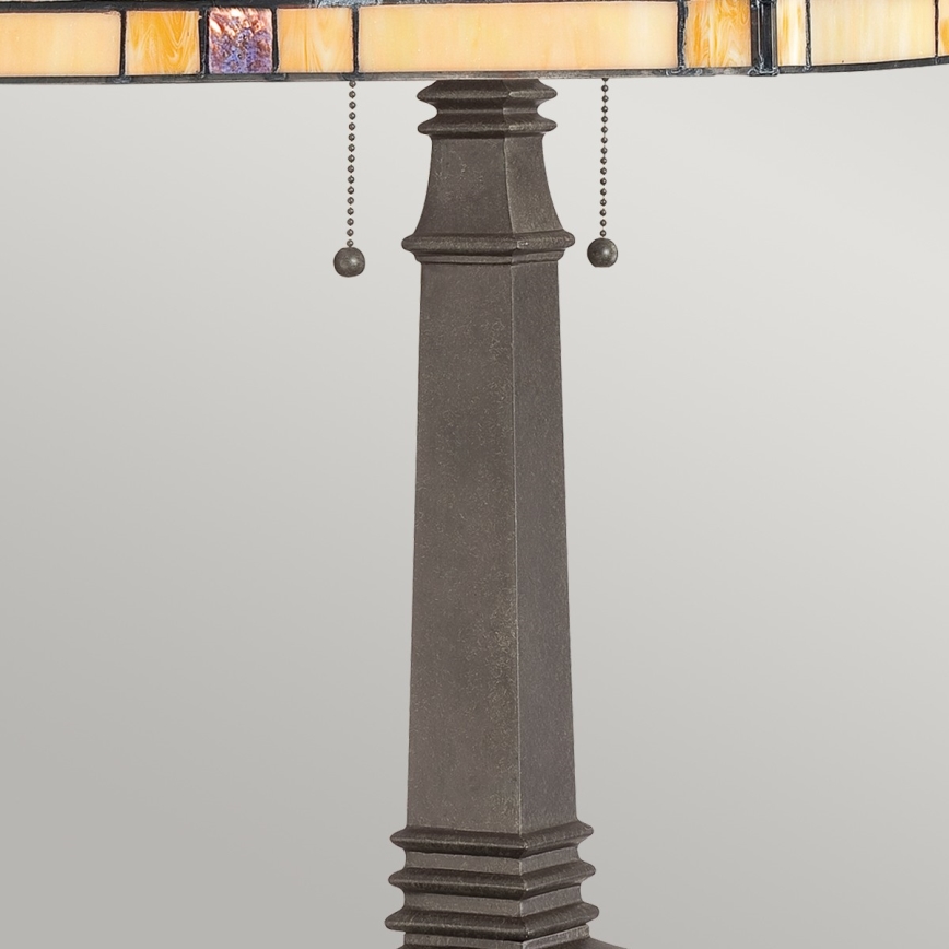 Stolová lampa s tienidlom z farebného skla stojí na tmavosivej kovovej nohe. Tienidlo je zdobené geometrickými vzormi. Noha má zúžený tvar a zakončenie v podobe ozdobných pásov.