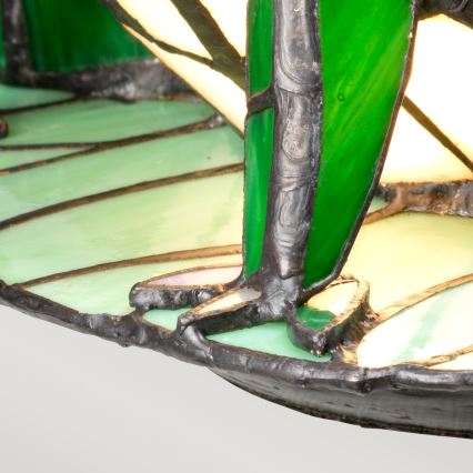Vitrážová lampa s motívom žaby stojí na plochej podložke. Nohy žaby sú z tmavého kovu, telo je zo zeleného a svetlozeleného skla. Svetlo presvitá cez sklo.