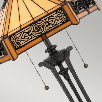 Stolová lampa s tienidlom z farebného skla, zdobeným ornamentálnym vzorom, stojí na trojnohom podstavci. Lampa je vybavená retiazkovým vypínačom.