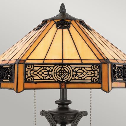 Stolová lampa s tienidlom z Tiffany skla, svetložltého odtieňa s hnedým kovovým lemovaním, stojí na tmavom kovovom podstavci. Tienidlo je zdobené ornamentálnymi detailmi. Osvetlenie je regulované retiazkou.