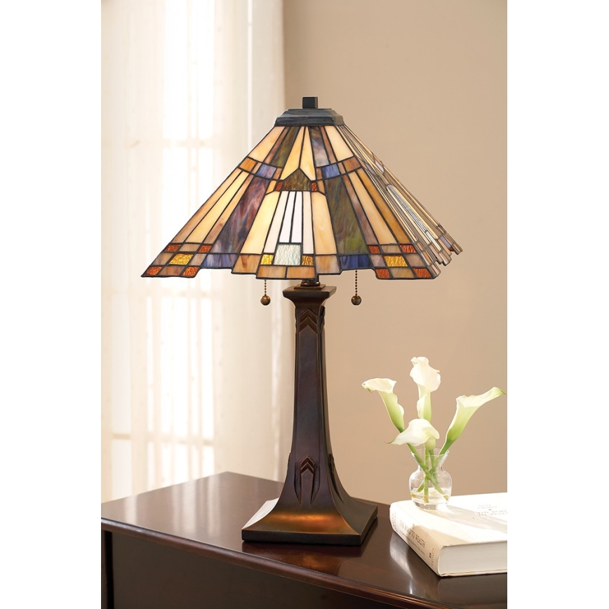 Stolová lampa so štvorcovou vitrážovou tienidlom v hnedo-modrých odtieňoch stojí na hnedom drevenom nábytku. Vedľa nej je váza s kvetmi a kniha.