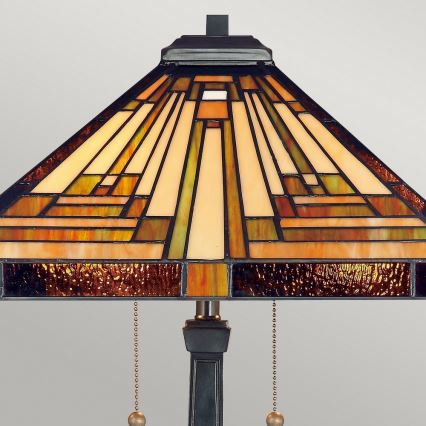Stolová lampa s tienidlom zo sklenených štvorcov v hnedých, béžových a zelených odtieňoch, stojí na tmavom podstavci. Zavesené sú na nej retiazky na zapínanie a vypínanie.