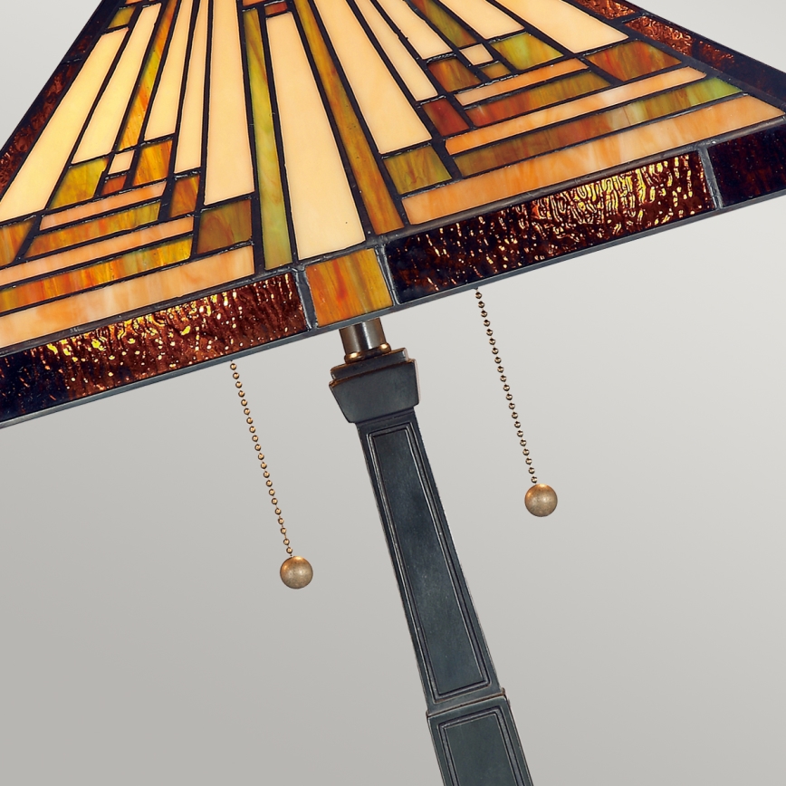 Stolová lampa s tienidlom z farebného skla v geometrickom dizajne, ovládaná retiazkami, stojí na tmavosivom podstavci.
