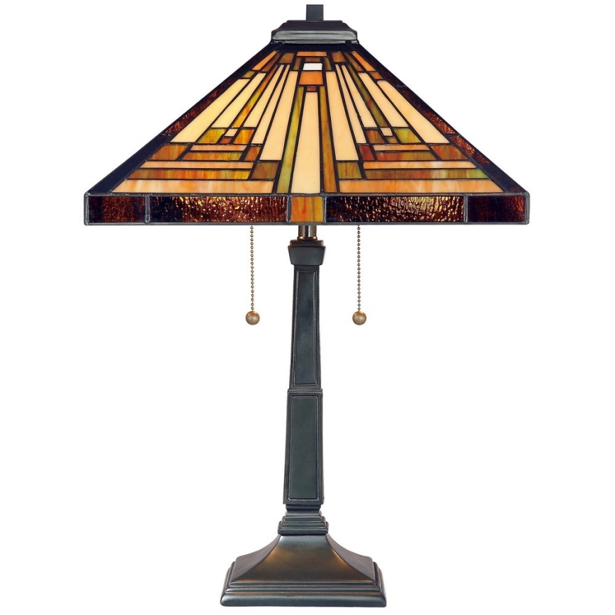 Stolová lampa s štvorcovou tienidlom z farebného skla s geometrickým vzorom osvetľuje priestor. Lampa má tmavý kovový podstavec a dva retiazkové vypínače.