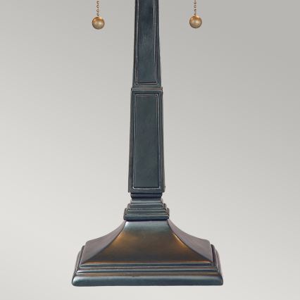 Stolová lampa s tmavosivou kovovou nohou v tvare štvorbokého hranola, zakončenou štvorcovou podstavou. Nohu zdobia dve retiazky s guličkami. Lampa stojí na svetlom pozadí.