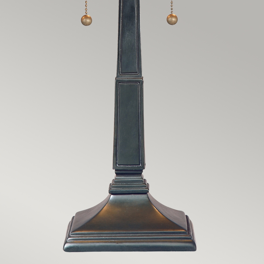 Stolová lampa s tmavosivou kovovou nohou v tvare štvorbokého hranola, zakončenou štvorcovou podstavou. Nohu zdobia dve retiazky s guličkami. Lampa stojí na svetlom pozadí.