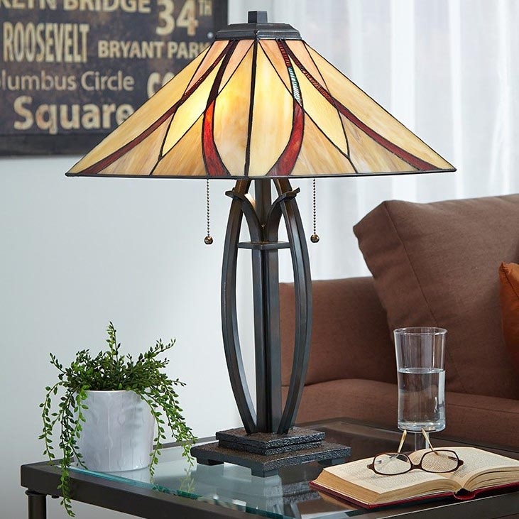 Stolová lampa s tienidlom z farebného skla stojí na odkladacích stolíku. Lampa má tmavohnedý kovový podstavec a tienidlo s béžovými, červenými a hnedými geometrickými vzormi. Na stolíku je tiež kvetináč s rastlinou, kniha a okuliare. Na stene za lampou je vidieť časť nápisu: ROOSEVELT, BRYANT PARK, Columbus Circle, Square.