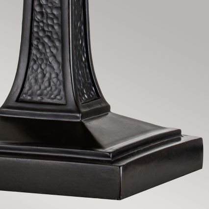 Stojan lampy stojí na svetlom pozadí. Je tmavej farby, s výraznou textúrou na strednej časti a štvorcovou základňou.