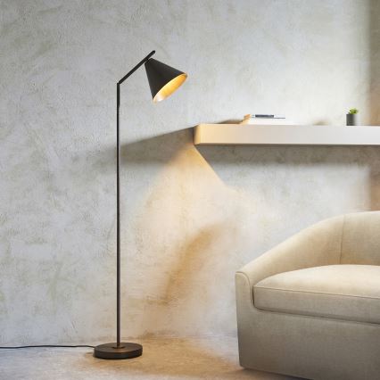 Stojacia lampa s kuželovým tienidlom svieti v miestnosti. Lampa je tmavosivá, tienidlo zvnútra zlaté. Nachádza sa pri svetlej sedačke a bielej poličke na stene z neomietnutej omietky.