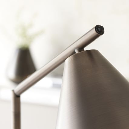 Stolová lampa s kužeľovitým tienidlom z matného kovu je umiestnená na stole. Rameno lampy je nastaviteľné. V pozadí je rozostrená váza s rastlinou.