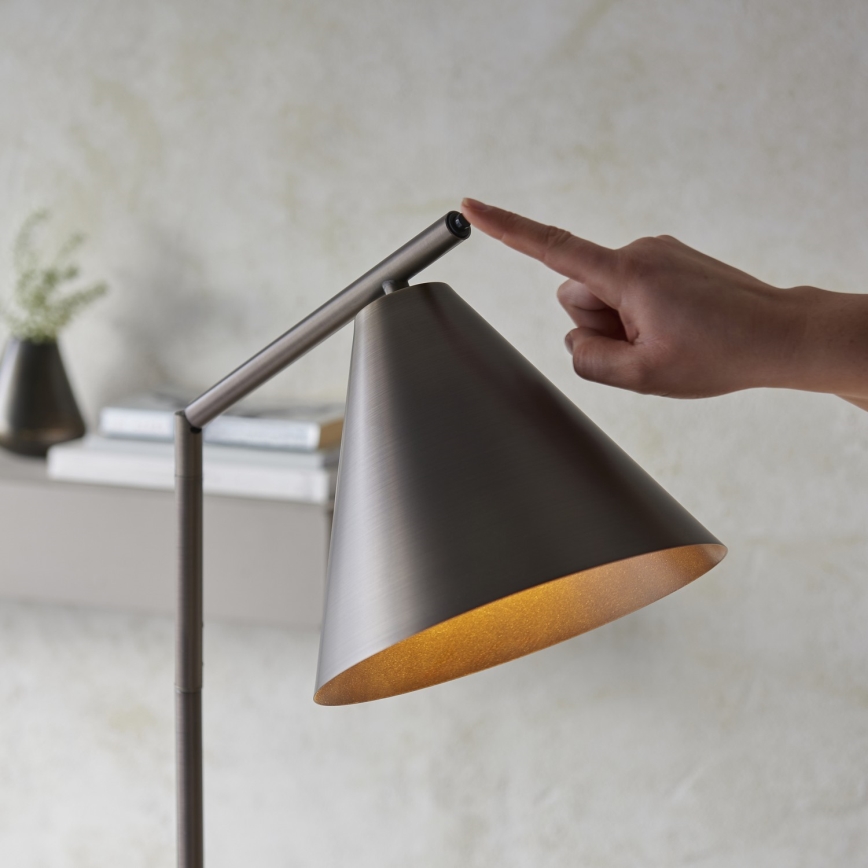 Stojacia lampa s kuželovitým tienidlom, sivého kovu, je zapínaná prstom. Lampa sa nachádza v interiéri, na pozadí je vidieť poličku s knihami a vázičku.
