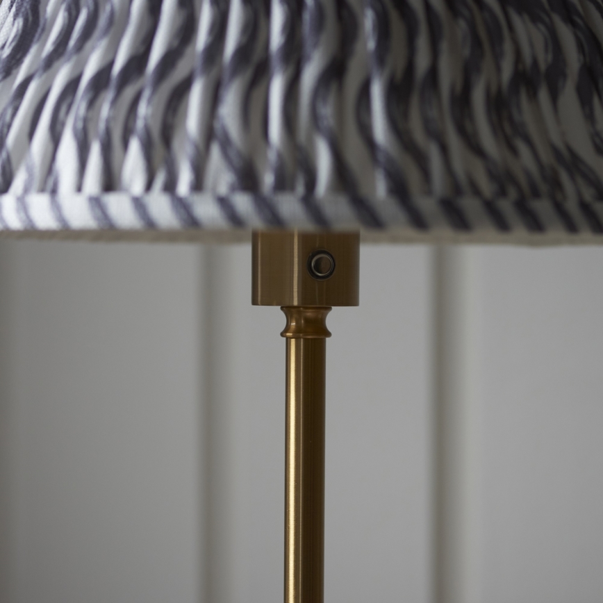 Stolová lampa s kovovou nohou a tienidlom s pruhovaným vzorom v sivej a bielej farbe má na stonke umiestnené vypínač. Lampa je v svetlom interiéri.