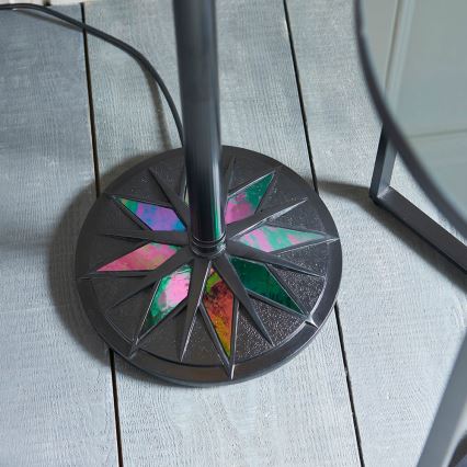 Stojaca lampa s čiernou kovovou nohou a okrúhlou podstavou. Podstava je zdobená farebnými, geometrickými výplňami. Lampa stojí na sivej drevenej podlahe.