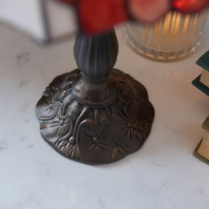 Stolová lampa s červeným tienidlom z farebného skla stojí na ozdobnej kovovej podstavci s reliéfnymi kvetmi. Podstava je tmavohnedá, lampa sa nachádza na svetlom mramorovom povrchu vedľa skleneného pohára a kníh.