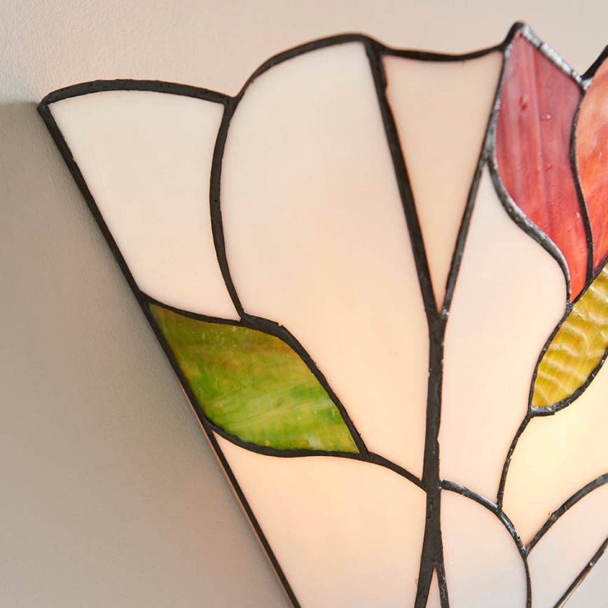 Nástenná lampa s kvetinovým motívom je pripevnená na stene. Je vyrobená z farebného skla v odtieňoch bielej, zelenej a ružovej, s čiernym kovovým lemovaním. Svieti.