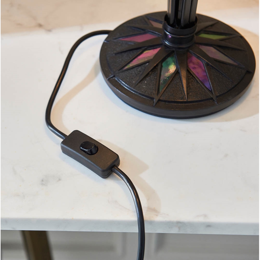 Stolová lampa s tmavohnedým podstavcom zdobeným farebnými sklenenými trojuholníkmi stojí na bielom povrchu. Je pripojená k sieti pomocou čierneho kábla so zapínacím vypínačom.