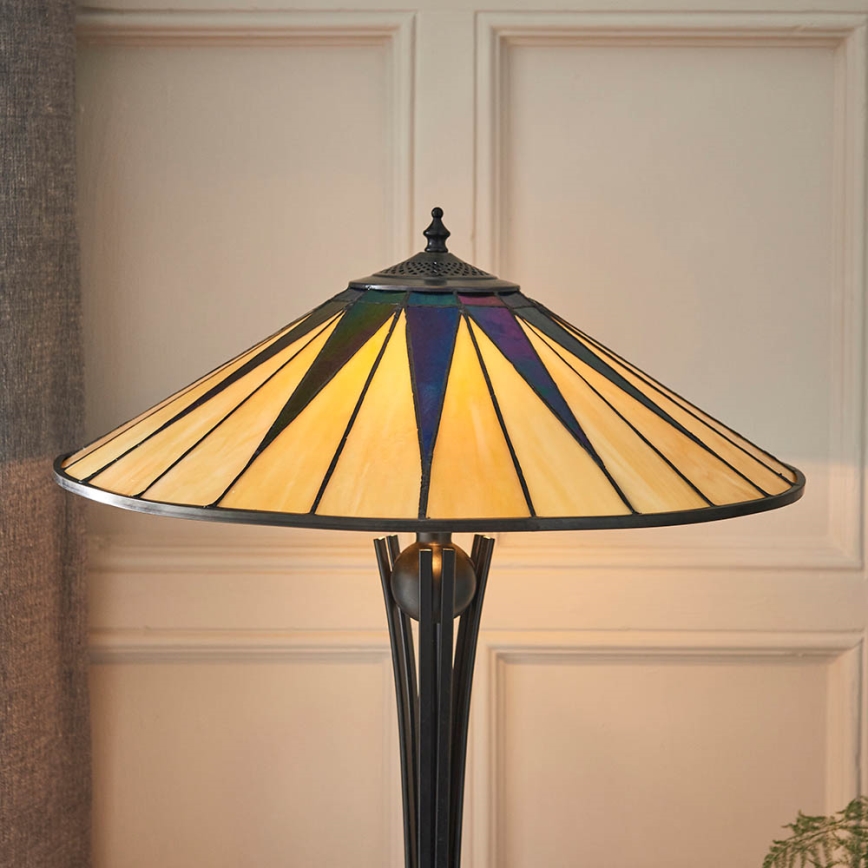 Stojacia lampa so širokým tienidlom zo skla s hnedými a modrými geometrickými vzormi na svetložltom pozadí. Lampa je osvetlená. Stojí pri svetlej stene s panelmi.