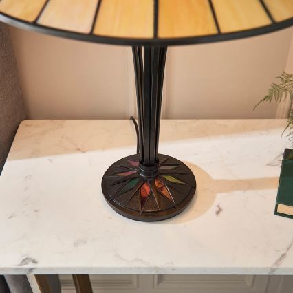 Stolová lampa s tienidlom zo svetlohnedého vitráža stojí na bielom mramorovom stolíku. Noha lampy je tmavá kovová s farebnými vitrážovými detailmi na podstavci.