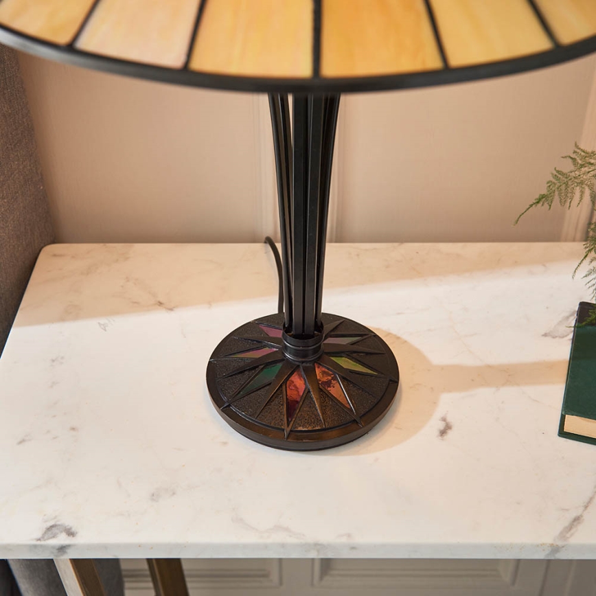 Stolová lampa s tienidlom zo svetlohnedého vitráža stojí na bielom mramorovom stolíku. Noha lampy je tmavá kovová s farebnými vitrážovými detailmi na podstavci.