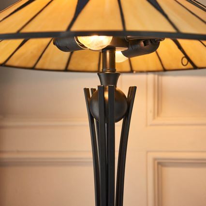 Stolová lampa s tienidlom z farebného skla svieti. Jej noha je z tmavého kovu, zdobená vertikálnymi prvkami. Lampa stojí na svetlom pozadí.