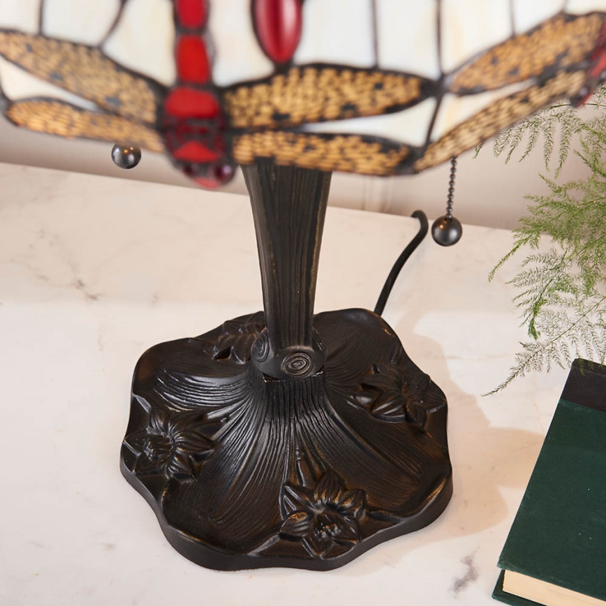 Stolová lampa s tienidlom zobrazujúcim vážky stojí na svetlom povrchu. Lampa má čierny kovový podstavec s kvetinovými motívmi a šnúrkový vypínač. V okolí je vidieť časť zelenej rastliny a knihu.