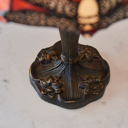 Stolová lampa s tienidlom z farebného skla stojí na tmavohnedej kovovej podstavci zdobenej kvetinovými motívmi. Podstavec má tvar rozprestretých lístkov. Lampa sa nachádza na svetlom povrchu.