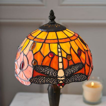 Stolová lampa s tienidlom z farebného skla s obrázkom vážky stojí na bielom stolíku. Vedľa lampy je sviečka a kniha.