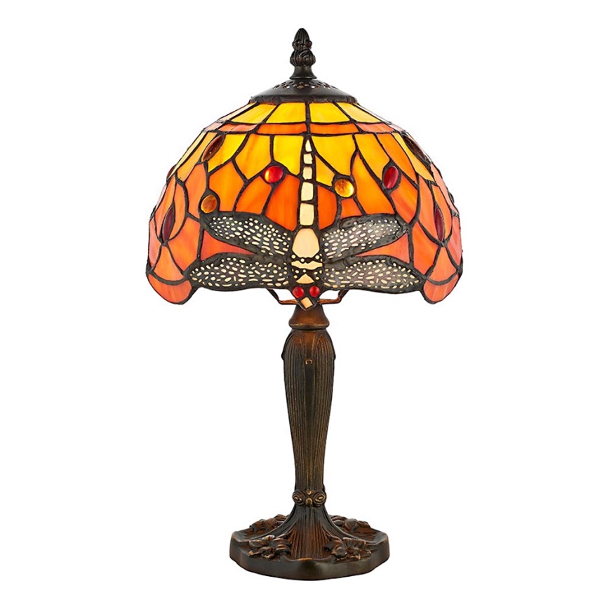 Stolová lampa s tienidlom z farebného skla zobrazuje vážku. Tienidlo je prevažne oranžové a žlté. Lampa stojí na stole vedľa sviečky.