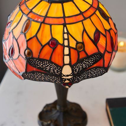 Stolová lampa s tienidlom z farebného skla zobrazuje vážku. Tienidlo je prevažne oranžové a žlté s červenými a hnedými detailmi. Lampa stojí na tmavom podstavci.