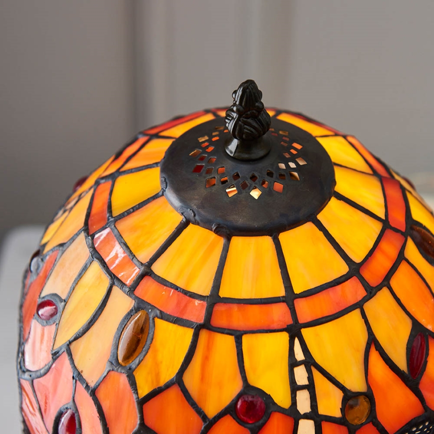 Stolová lampa s tienidlom z farebného skla v odtieňoch oranžovej a červenej. Tienidlo je zdobené geometrickým vzorom. Vrch tienidla je tmavo hnedý s otvormi.