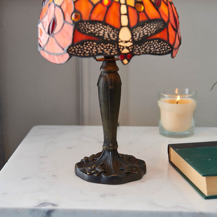 Stolová lampa s tienidlom z farebného skla s motívom vážky stojí na mramorovom stole. Vedľa nej je zapálená sviečka a kniha.
