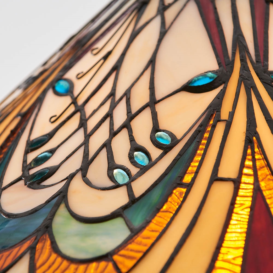 Tienidlo lampy s vitrážovým vzorom zobrazuje krivky a línie v béžovej, oranžovej a modrej farbe. Tmavé špáry oddeľujú farebné sklenené časti. Modré kamienky sú vsadené do vzoru.