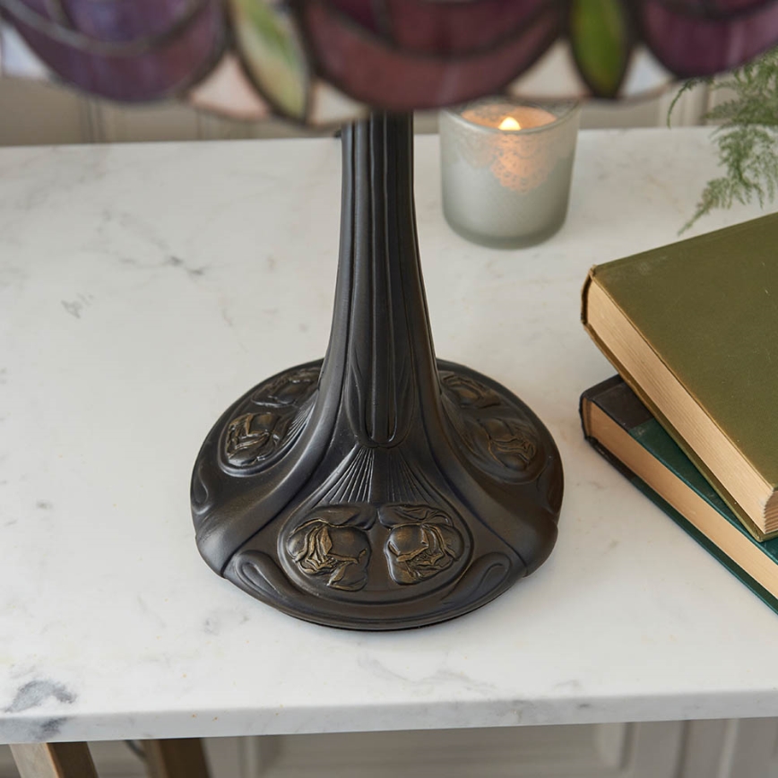 Stolová lampa s tienidlom z farebného skla stojí na mramorovom stole. Jej kovová základňa je zdobená reliéfnymi kvetmi. Na stole sa nachádza aj sviečka a knihy.