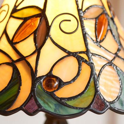 Tiffany lampa s tienidlom z farebného skla zobrazuje ornament listov a spirál. Svetlo presvitá cez sklo. Tmavý kovový podstavec.
