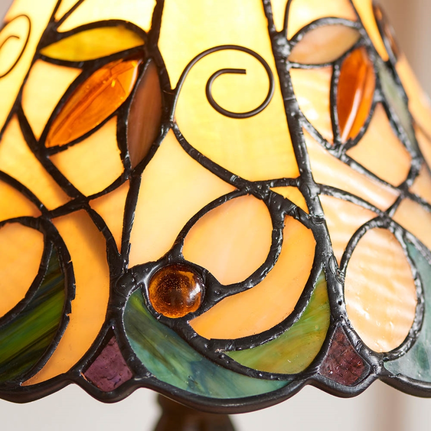 Tiffany lampa s tienidlom z farebného skla zobrazuje ornament listov a spirál. Svetlo presvitá cez sklo. Tmavý kovový podstavec.