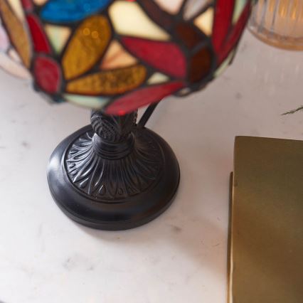 Stolová lampa s tienidlom z farebného skla stojí na mramorovom povrchu vedľa knihy. Tienidlo je zdobené rôznofarebnými listami. Noha lampy je tmavá, kovová.