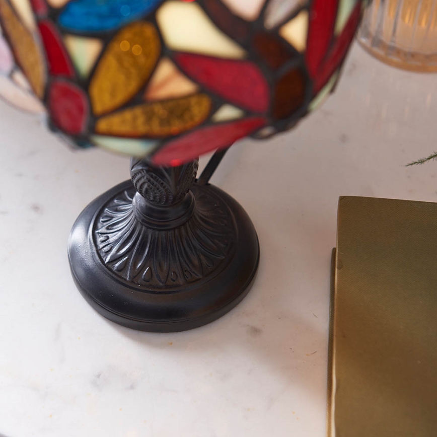 Stolová lampa s tienidlom z farebného skla stojí na mramorovom povrchu vedľa knihy. Tienidlo je zdobené rôznofarebnými listami. Noha lampy je tmavá, kovová.