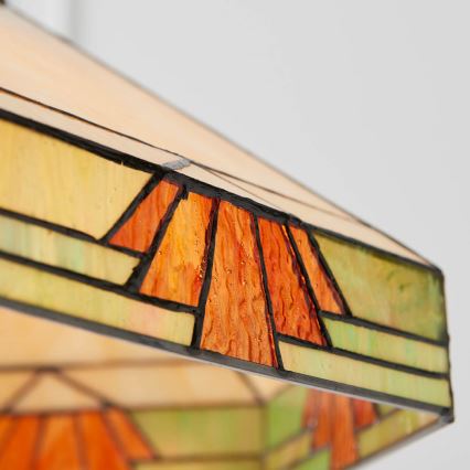 Svietidlo s tienidlom zo skla s geometrickým vzorom v oranžovej a zelenej farbe je zobrazené zblízka. Tienidlo je štvorcové s fasetami. Na detailnom zábere je vidno textúru skla.