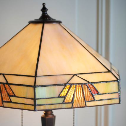 Stolová lampa s tienidlom z Tiffany skla svieti. Tienidlo má geometrický vzor v odtieňoch béžovej, zelenej a oranžovej. Lampa stojí na tmavom drevenom podstavci.