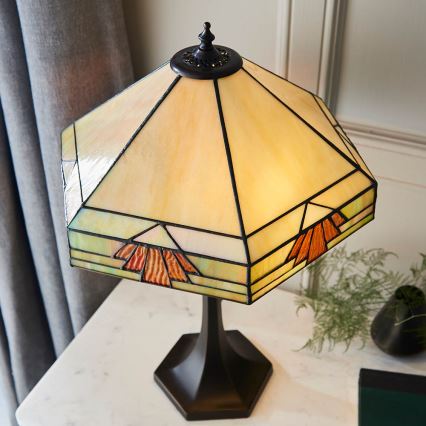 Stolová lampa s osmibokou tienidlom zo skla s hnedými a zelenými geometrickými motívmi svieti na bielom stolíku. Lampa má tmavohnedý kovový podstavec.
