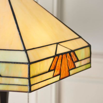 Stolová lampa s pyramídovým tienidlom z farebného skla. Tienidlo je zložené z béžových, zelenkavých a oranžových sklenených častí s tmavými kovovými obrubami. Lampa je umiestnená na svetlom pozadí.