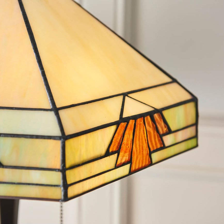 Stolová lampa s pyramídovým tienidlom z farebného skla. Tienidlo je zložené z béžových, zelenkavých a oranžových sklenených častí s tmavými kovovými obrubami. Lampa je umiestnená na svetlom pozadí.