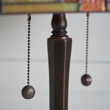 Stolová lampa s bronzovou nohou a tienidlom má dva kovové šnúrky s guľôčkami na zapínanie a vypínanie. Jeden z guľôčok má vyrytý nečitateľný text. Lampa je umiestnená na svetlom pozadí.